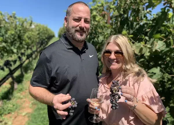 Smiling couple enjoying a wine tour in a Virginia vineyard, holding grapes and glasses of wine during the Middleburg Experience.